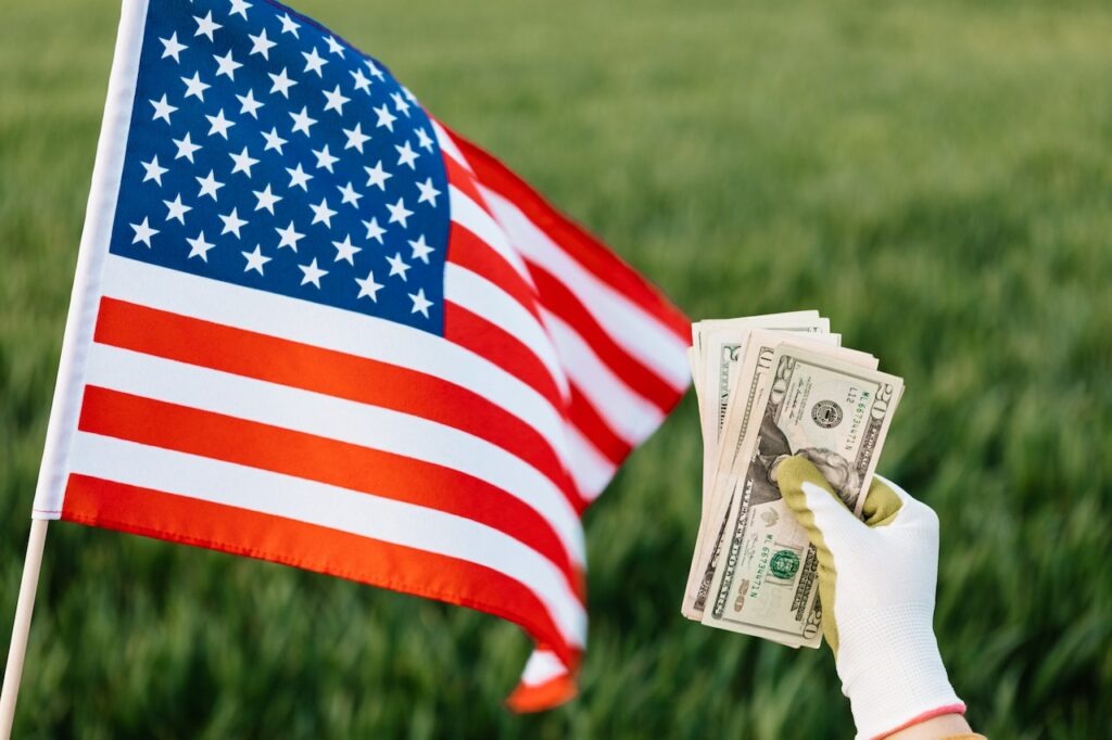 American flag and crop unrecognizable person with pile of dollar from selling land on Labor day