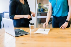 Two women standing over a table with lap top and document with pen in one woman's hand to sign next to a cup of take away coffee