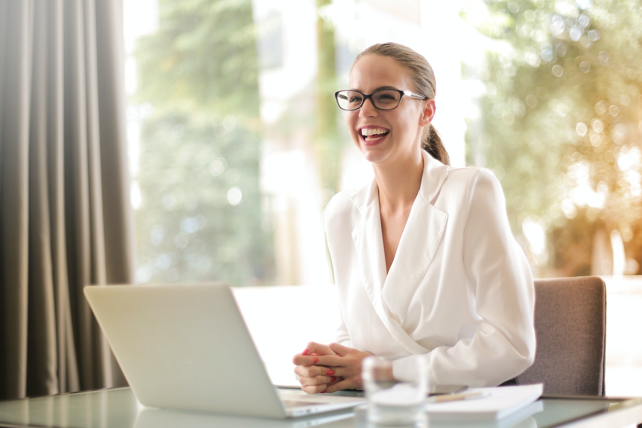 Young woman with glasses sitting on desk with opened laptop next to a glass of water smiling about the process of buying land with cash